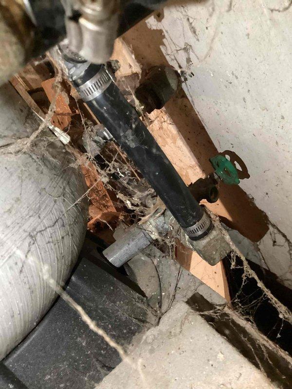 Responded to water softener leak complaint and performed on-site inspection of residential water treatment system. Upon arrival, confirmed active leak from water softener unit with cream-colored fiberglass tank and rust-orange top cap mounted on basement wall. Inspected entire plumbing assembly including copper piping, control equipment, motor assembly, and valve connections. Documented deteriorated wooden support structures showing water damage, rot, and mold growth in crawl space area affecting adjacent plumbing infrastructure. Photographed condition of equipment, piping connections with metal hose clamps, green valve handle assembly, and overall installation including visible corrosion on older metal components. Assessed water softener system functionality and identified source of leak requiring repair or replacement recommendations.