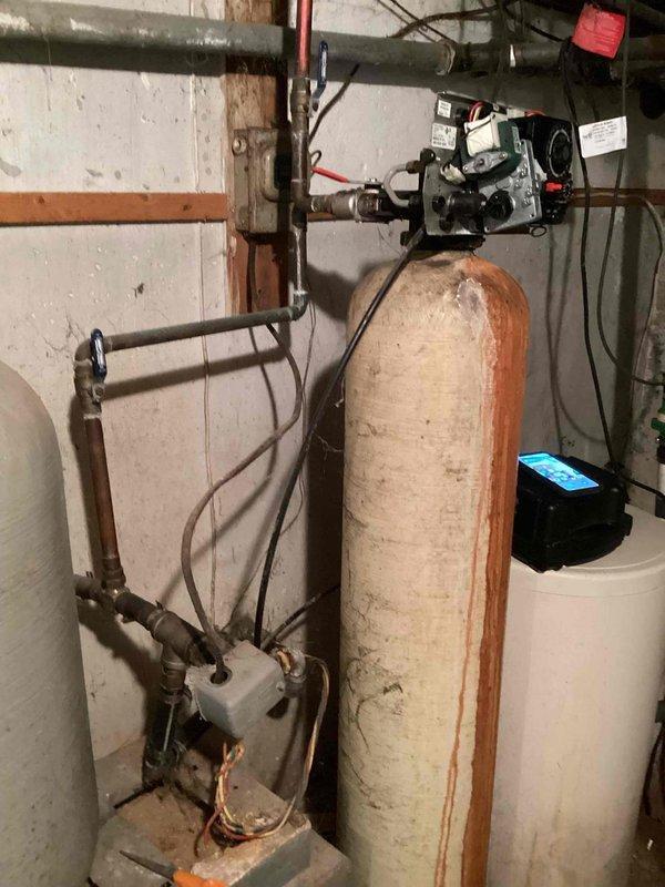 Responded to water softener leak complaint and performed on-site inspection of residential water treatment system. Upon arrival, confirmed active leak from water softener unit with cream-colored fiberglass tank and rust-orange top cap mounted on basement wall. Inspected entire plumbing assembly including copper piping, control equipment, motor assembly, and valve connections. Documented deteriorated wooden support structures showing water damage, rot, and mold growth in crawl space area affecting adjacent plumbing infrastructure. Photographed condition of equipment, piping connections with metal hose clamps, green valve handle assembly, and overall installation including visible corrosion on older metal components. Assessed water softener system functionality and identified source of leak requiring repair or replacement recommendations.