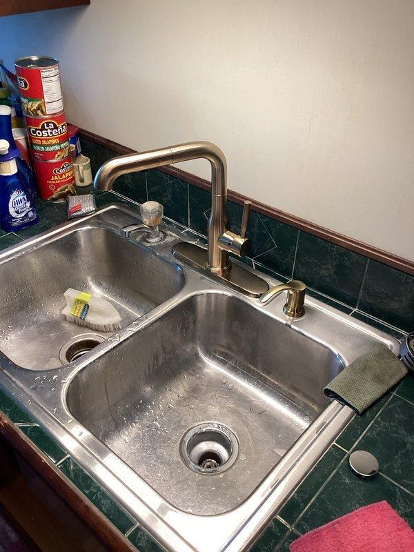 Replaced kitchen faucet with homeowner-provided brushed nickel pull-down model featuring high-arc spout design. Installed new faucet assembly on stainless steel double-basin sink with dark green tile countertop, ensuring proper mounting and connection to existing water supply lines and drain configuration.