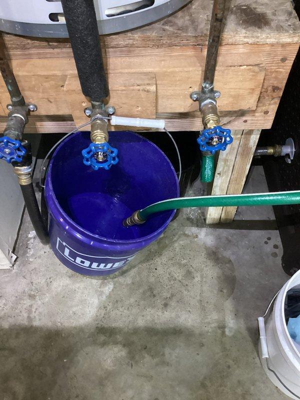 Responded to callback for dripping valve in garage following recent water heater replacement. Upon arrival, inspected gate valve with blue handle connected to water supply system near pressure tank. Valve exhibited continuous dripping after being cycled open and closed. Assessed brass fittings, connections, and valve assembly for potential seat damage or debris causing improper seal. Documented plumbing configuration including pressure tank, gate valves, drain setup with hose connection to bucket, and associated piping. Photographed installation to record current condition and identify source of leak for repair recommendations.